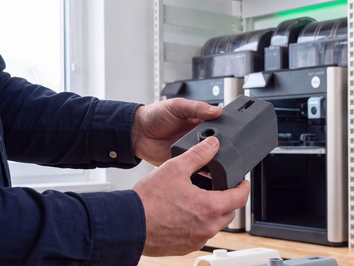 Ruce držící 3D tištěný prototyp dílu před řadou tiskáren v dílně